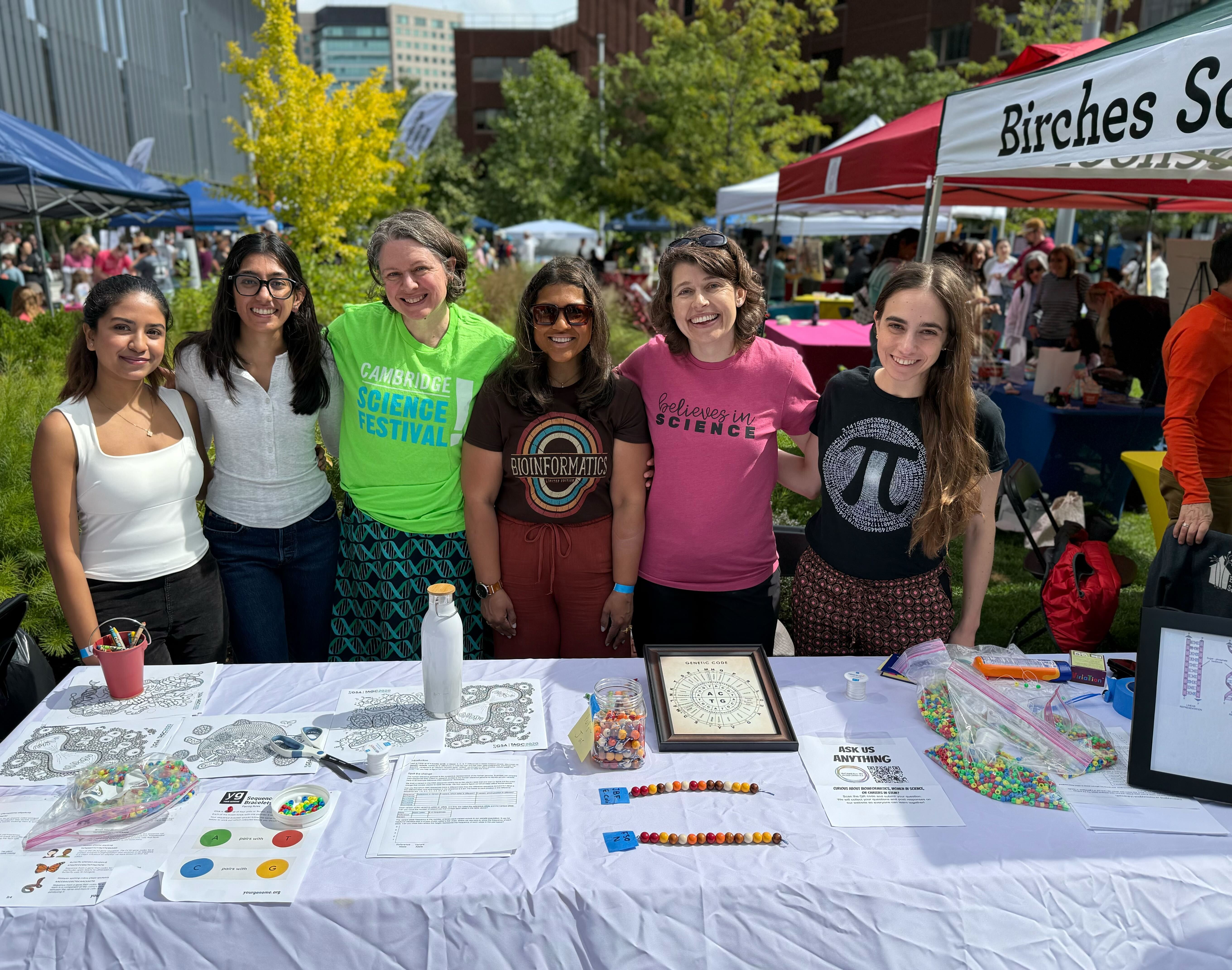 Six women at Cambridge Science Festival booth with kids activity table. Shirts say Cambridge Science Festival, Bioinformatics with rainbow, Believes in Science, and pi symbol.